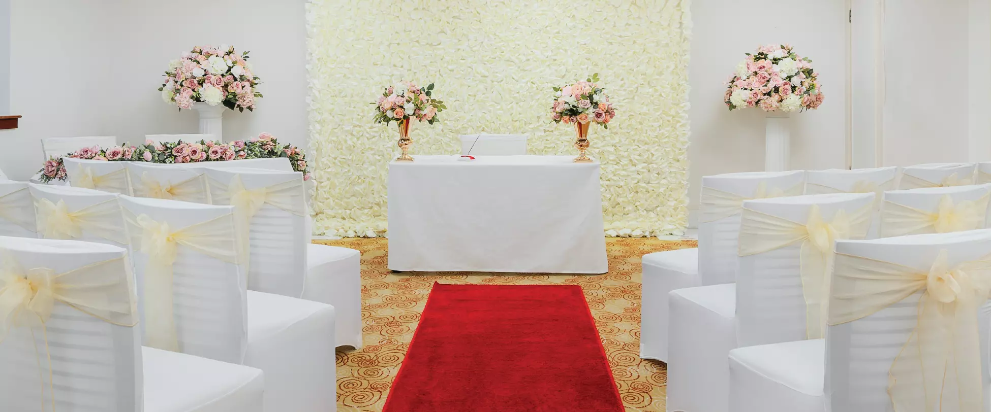 A room set up for a wedding ceremony. A table at the front is covered in white and has bouquets of pink flowers to either side. There's a wall of yellow flowers behind the table and a red carpet leading up. Either side of the carpet are chairs with ribbon.