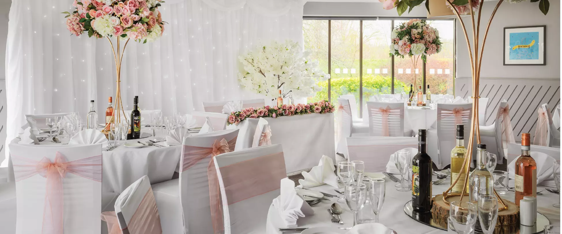 A room set up for a wedding breakfast with tables and chairs covered in white, pink ribbons and flowers..